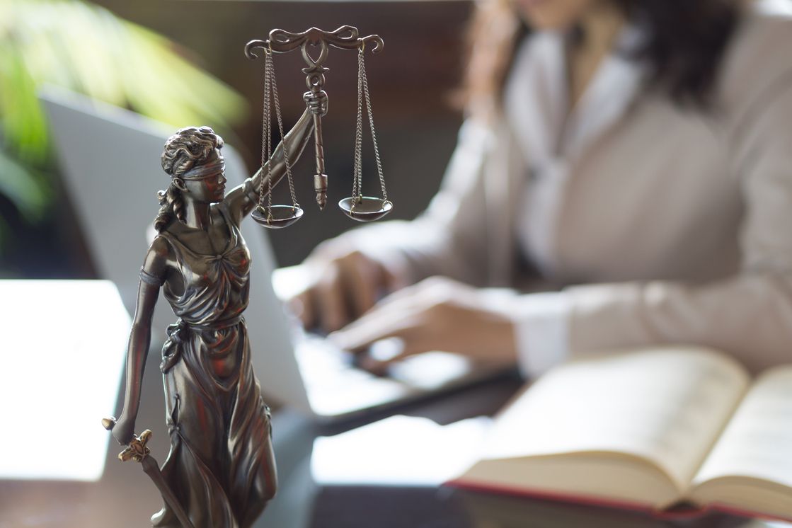 Eine Justitia steht auf dem Schreibtisch einer im Hintergrund arbeitenden Anwältin